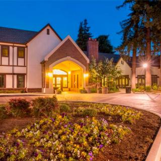 A charming house with a beautifully lit entrance, surrounded by gardens and trees during the evening.