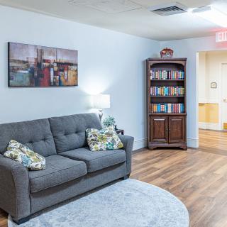 A cozy living room featuring a gray sofa with decorative pillows, a wooden bookshelf filled with books, and a framed wall art piece. The space is warmly lit by a lamp on the side.
