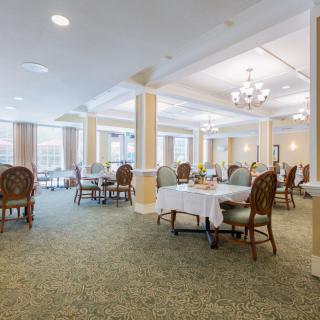 A spacious and elegantly decorated dining room with tables and chairs arranged neatly, featuring chandeliers and large windows providing natural light.