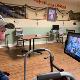 A smiling senior in a nursing home participates in a video call, surrounded by Halloween decorations.