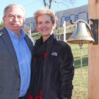 A smiling couple poses outdoors near a bell on a sunny day.