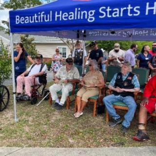 A community gathering under a tent with a sign that reads 'Beautiful Healing Starts Here'.