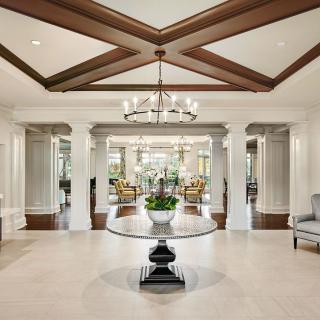 A luxurious foyer featuring elegant columns, a central table with a floral arrangement, and an ornate chandelier hanging from a coffered ceiling.