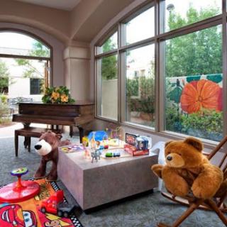 A cozy playroom filled with soft toys and a grand piano, bathed in natural light from large windows.
