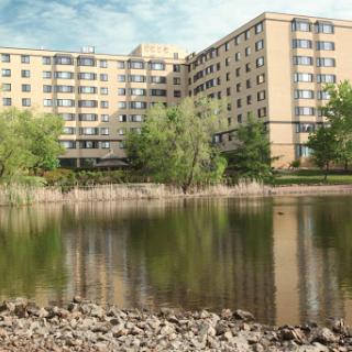 A serene view of an apartment building reflected in a calm lake, surrounded by greenery and a rocky shoreline.