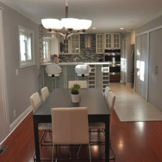 A modern dining room with a view of the kitchen, featuring a table, chairs, and a chandelier on hardwood flooring.