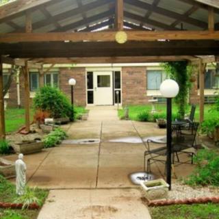 A serene garden path leading to a brick building, featuring outdoor seating and decorative lamp posts under a covered patio.