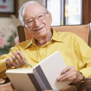 An elderly man sitting in a chair and smiling while holding an open book.