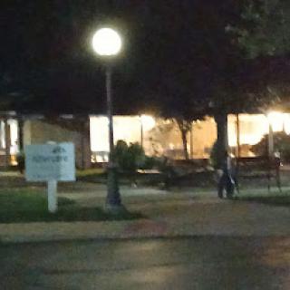 A dimly lit park scene at night with a lamp illuminating the pathway and a building in the background.