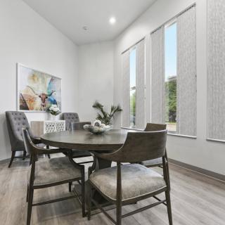 A modern dining room with a round wooden table, surrounded by upholstered chairs and large windows allowing natural light. Minimalist decor includes artwork and an indoor plant.