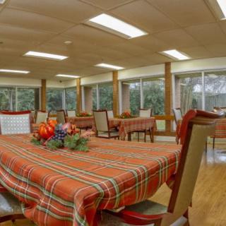 A cozy dining room with tables set in plaid tablecloths and decorated with floral arrangements, illuminated by natural light through large windows.