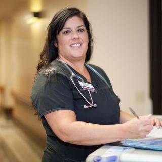 A nurse in scrubs with a stethoscope, standing in a hospital hallway and writing notes.