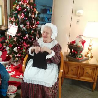 An elderly woman in traditional attire enjoys knitting by the Christmas tree.