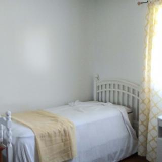 A minimalistic bedroom with a single bed and white bedding, softly illuminated by natural light from a window.