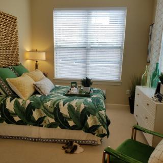 A cozy bedroom with green and natural tones, featuring a patterned bedspread, plants, and soft lighting.
