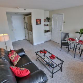 A cozy apartment living room featuring a black sofa with red cushions, a glass coffee table, and an adjacent dining area.