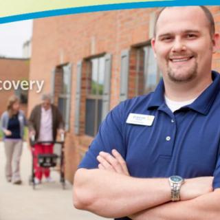 A smiling staff member stands confidently outside a healthcare facility, with people strolling in the background.