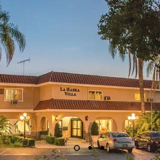 A charming villa-style hotel building illuminated in the evening, surrounded by palm trees and parked cars.