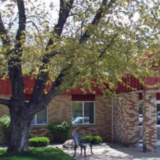 A brick building with a large tree in front, accompanied by outdoor seating on a pathway.