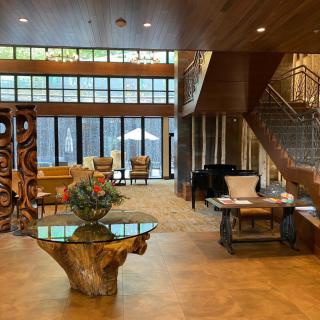 A spacious and modern lobby featuring rustic wooden furniture, a staircase, and large glass windows allowing natural light to fill the space.