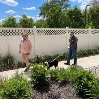 A couple walking their dog along a sunny sidewalk next to a white fence.