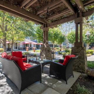 A cozy outdoor patio with wicker furniture and red cushions, under a wooden gazebo overlooking a garden.