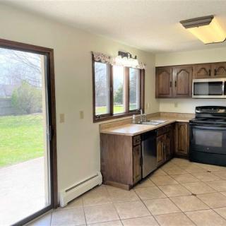 A bright kitchen with wooden cabinets, tile flooring, and modern appliances, featuring a window and a sliding glass door opening to the outside.