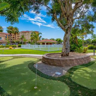 A scenic view of a putting green with a large tree and nearby resort buildings under a clear blue sky.