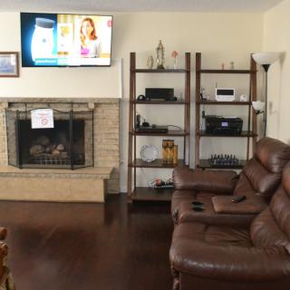 A cozy living room with a leather couch, fireplace, and a TV mounted above. Shelves are filled with various electronics and decor items.