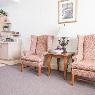 A cozy waiting area with two patterned chairs flanking a side table and lamp, beneath a framed artwork.