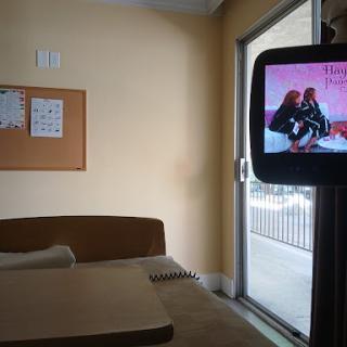 A view of a hospital room displaying a bed and a television showing content.