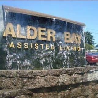 A sign for Alder Bay Assisted Living located outdoors, with a water feature in the background.