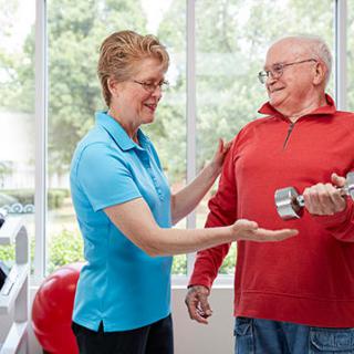 A senior man lifting a dumbbell with the assistance of a trainer in a well-lit room.