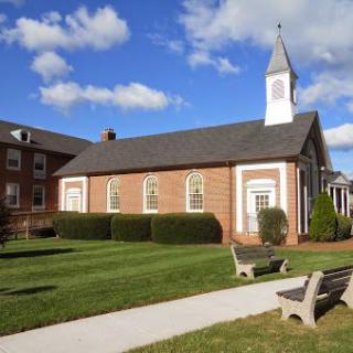 A small brick church with a steeple under a partly cloudy sky, surrounded by grass and benches.