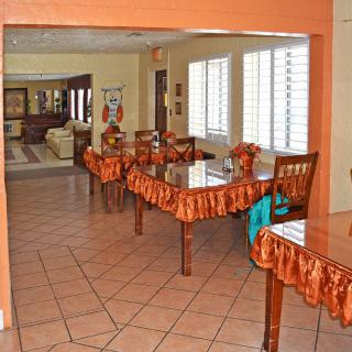 A cozy dining area with several wooden tables and chairs, decorated with orange tablecloths, set on a tiled floor with natural light streaming in through the windows.