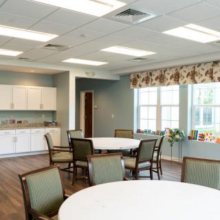 A bright and spacious dining area with round tables and chairs, featuring a kitchenette in the background.