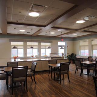 A spacious dining area with wooden floors, tables, and chairs, featuring large windows and ceiling lights.