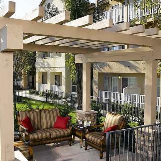 A cozy patio with a pergola, featuring cushioned chairs and surrounded by a residential complex with lush greenery.