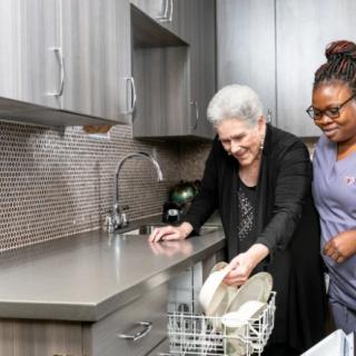 An elderly person and a caregiver loading dishes into a dishwasher in the kitchen.