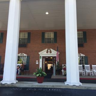 A brick building with large white columns and a porch featuring rocking chairs.