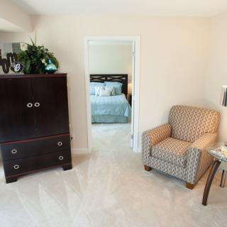 A cozy living area with a patterned armchair and a dark cabinet, leading into a bedroom.