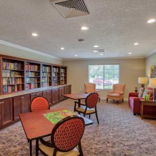 A cozy library room with bookshelves, tables, and chairs, featuring a large window providing natural light.