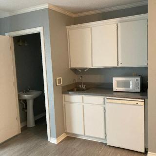 A compact kitchenette with white cabinets, a microwave, and a small refrigerator adjacent to an open bathroom door.