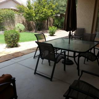 A cozy backyard patio featuring a glass table with chairs and an umbrella, overlooking a lush garden.
