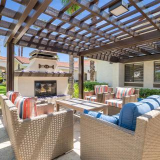 A sunny outdoor patio featuring comfortable furniture with colorful cushions, a fireplace, and a pergola.