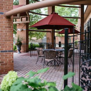 A cozy patio with tables, chairs, and a red umbrella set against a backdrop of brickwork and greenery.