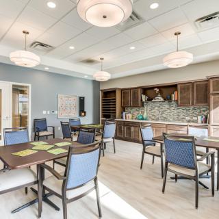 A modern dining area with neatly arranged tables and chairs, featuring overhead lighting and a kitchenette in the background.