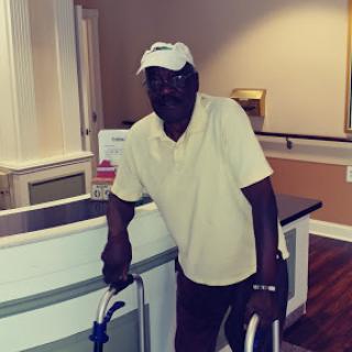 An older man using a walking aid stands by a reception area, dressed casually in a yellow shirt and white cap.