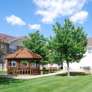 A serene outdoor area featuring a wooden gazebo, surrounded by lush green grass and trees, under a bright blue sky with scattered clouds.