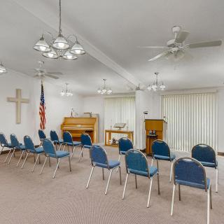 An interior view of a chapel with rows of blue chairs, a piano, a podium, and religious symbols.